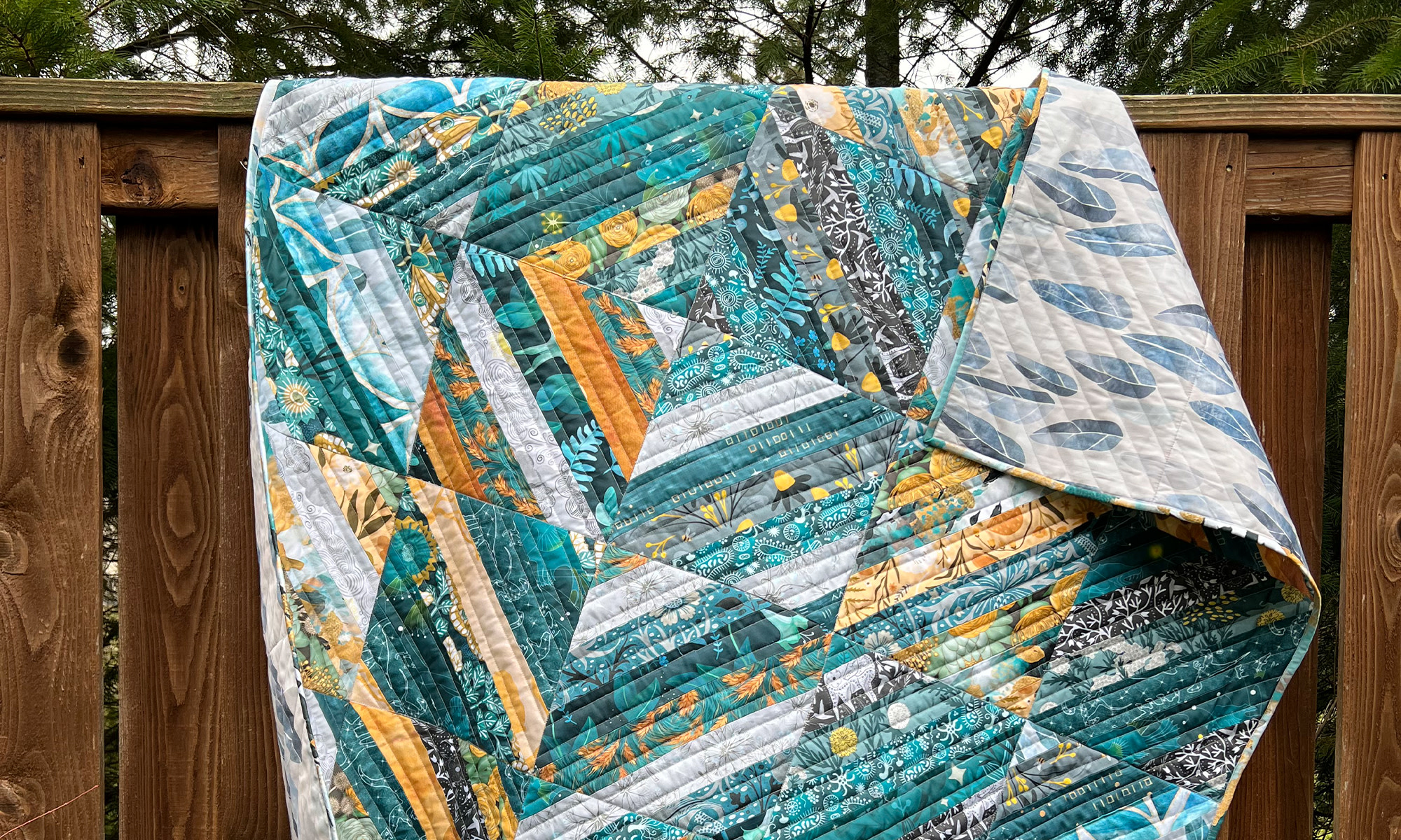 A quilt made of teal, yellow and white Spoonflower fabrics hanging on a fence.
